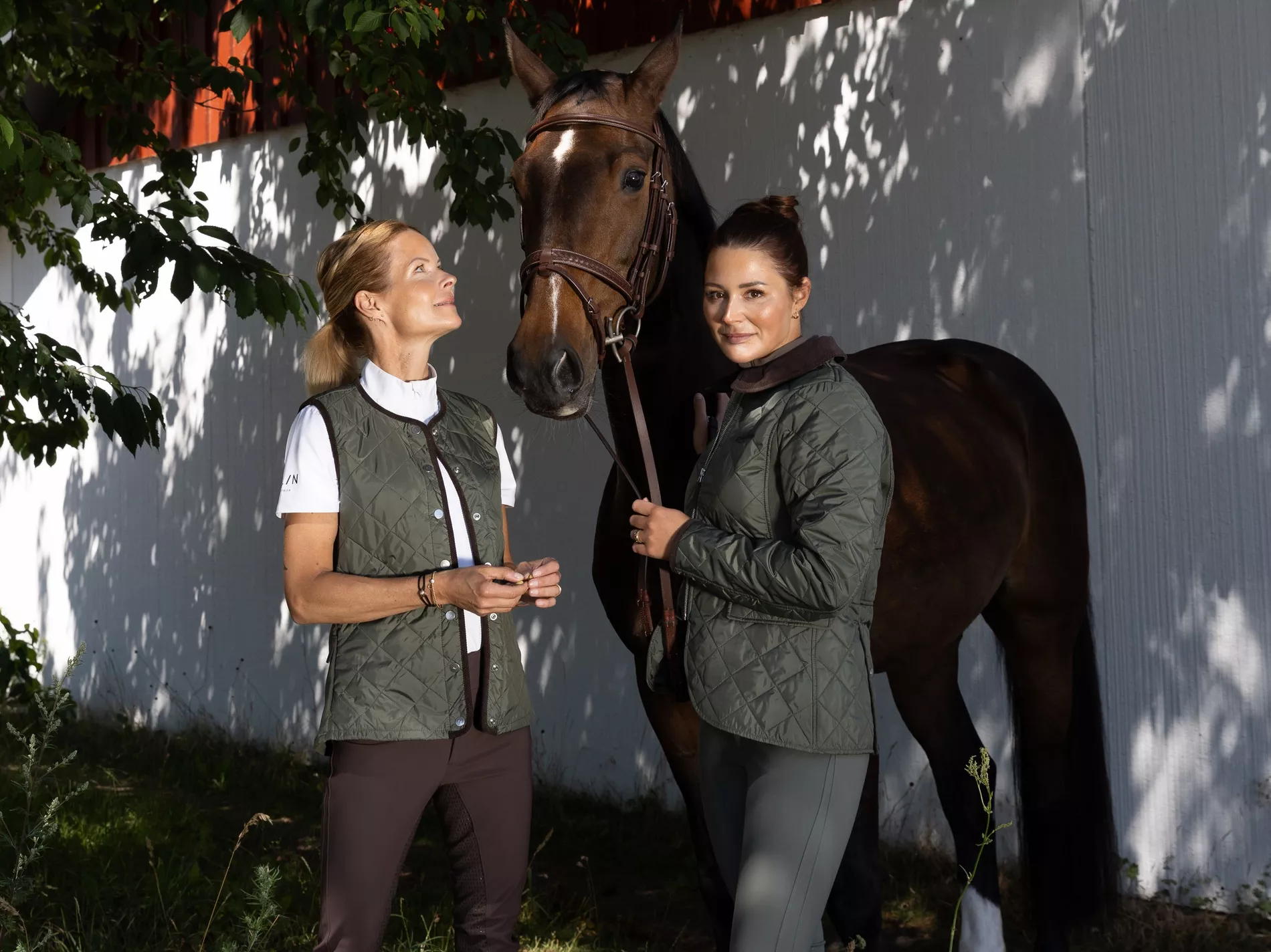 Two women in olive quilted equestrian outerwear standing with a bay horse in dappled sunlight